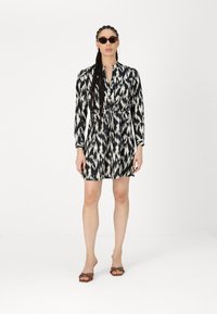 Schwarz-weißes Kleid mit Ikat-Muster, langen Ärmeln, Bund mit Bindegürtel und Knopfleiste. Getragen mit Sonnenbrille und braunen Absatzsandalen.