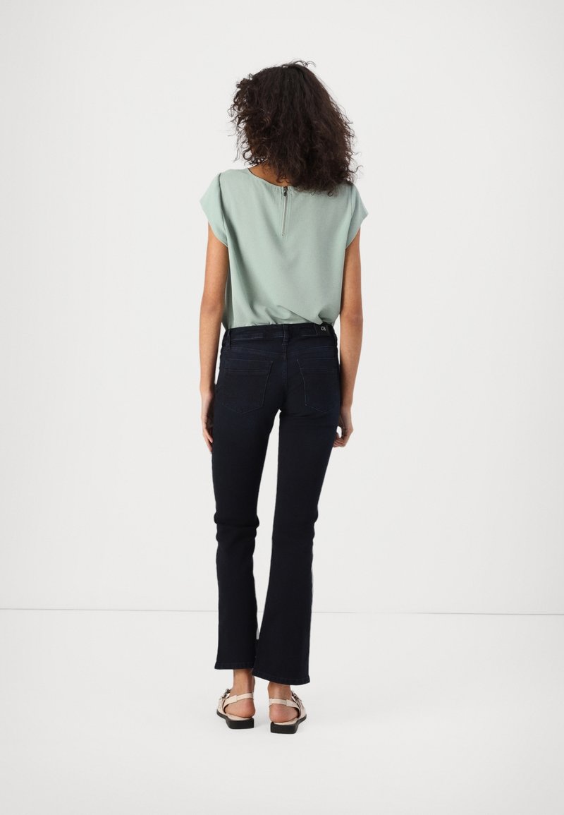 Femme aux cheveux bouclés portant un haut vert clair à manches courtes, un jean bleu foncé et des sandales, debout de dos sur un fond blanc.