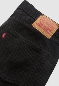 Zwarte corduroy Levi's spijkerbroek met zichtbare achterzak, rood Levi's-label en een lichtbruine leren patch met rood logo en tekst.