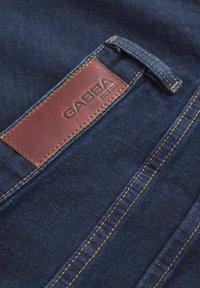Temno modra jeans tkanina z rumenim šivanjem; na hrbtni strani ima rjav usnjen oznako z odtisnjenim napisom "GABBA" in "SINCE 1983".