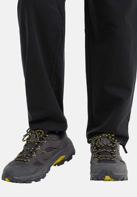 Graue Wanderschuhe mit gelben Akzenten und schwarzem Netzmaterial. Lebhafte gelbe Schnürsenkel und strukturierte Gummisohlen für besseren Halt. Kombiniert mit schwarzen Hosen.