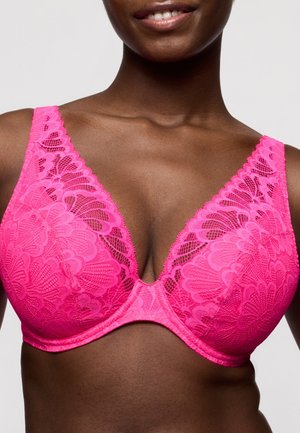 Vrouw die een felroze kanten beha draagt met golvende randen en bloemmotieven, licht glimlachend tegen een effen achtergrond.