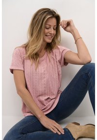 Haut rose clair à manches courtes avec un design à œillets et un col côtelé, associé à un jean sombre et des bottines beiges.