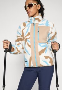 Donna che indossa una giacca in pile a fantasia, pantaloni navy, occhiali da sole beige, mentre tiene in mano dei bastoni da trekking neri, girata leggermente verso destra su uno sfondo semplice.