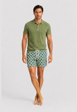 Ein Mann läuft barfuß und trägt ein grünes Poloshirt mit kurzen Ärmeln sowie grüne gemusterte Shorts mit Kordelzug vor einem einfarbigen Hintergrund.