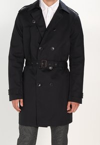 Svart dubbelknäppt trench coat med bälte, stora knappar, axelklappar och en skräddarsydd passform. Bärs över en vit skjorta och mönstrade byxor.