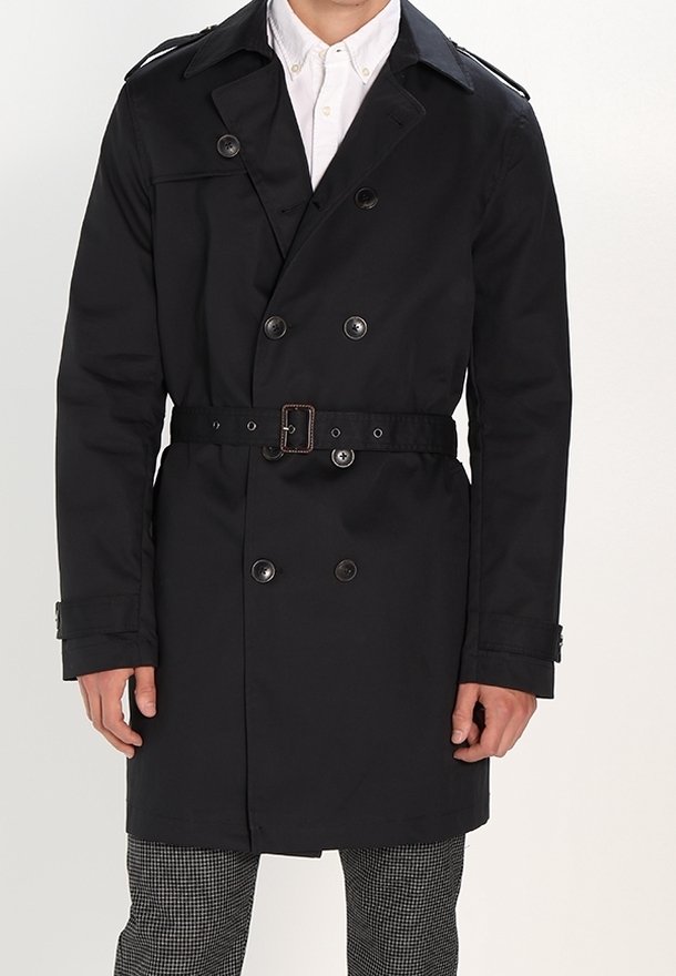 Svart dubbelknäppt trench coat med bälte, stora knappar, axelklappar och en skräddarsydd passform. Bärs över en vit skjorta och mönstrade byxor.