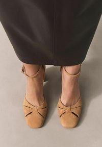 Suedeabsatzschuhe in Tan mit ausgeschnittenen Mustern, ausgestattet mit einer abgerundeten Zehenpartie und einem Knöchelriemen. Der Absatz ist etwa 5 cm hoch.