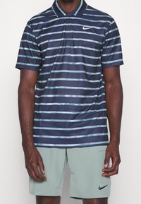 Polo shirt marine avec des rayures aqua claires, manches courtes et un col. Présente un logo Nike sur la poitrine. Texture de tissu respirant.