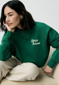 Grøn sweatshirt med ribbede manchetter og halsudskæring, der har "Rockin' Around" broderet i cremefarve. Modellen har beige bukser på og sidder på en lys overflade.
