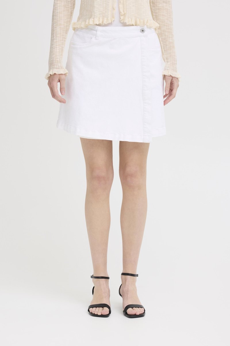 Femme portant une jupe portefeuille blanche, un haut beige à manches longues et des sandales noires à bride à la cheville, debout devant un fond uni.