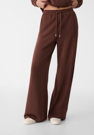 Pantaloni da jogging larghi di colore marrone, realizzati in tessuto morbido, con vita elastica, coulisse e tasche laterali. Texture liscia, vestibilità comoda.