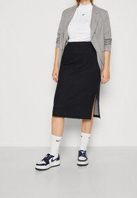 Harmaa bleiseri, valkoinen t-paita Nike-logolla, musta viivoitettu hame, jossa on sivuhalkio, ja tummansiniset sneakerit valkoisilla yksityiskohdilla ja Nike-logolla.