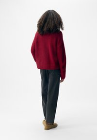 Czerwony, fakturowany, oversize sweter z opadającymi ramionami, w połączeniu z ciemnoszarymi spodniami i tanowymi butami wsuwanymi, widoczny z tyłu.