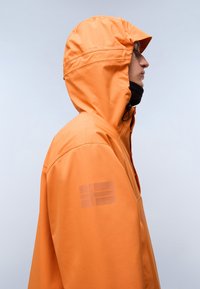 Person iført en orange hættejakke med en svag flaglogo på ærmet, vendt mod højre mod en ensfarvet lys baggrund.