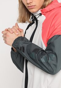 Coupe-vent avec capuche rouge et accents noirs, doté d'un corps léger gris et d'un motif de logo rayé noir sur les manches.