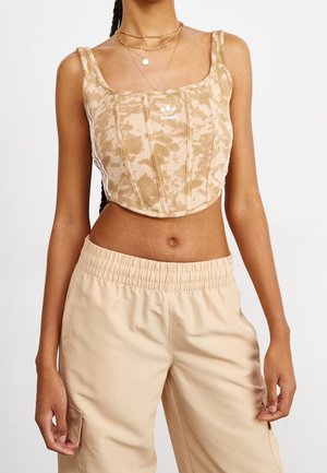 Femme portant un crop top beige à motifs Adidas et un pantalon beige clair taille élastique, avec des colliers dorés en couches.