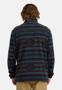 Fleece pullover met donkerblauwe, kastanjebruine en bruine strepen. Heeft een hoge kraag en lange mouwen. De textuur lijkt zacht en warm.