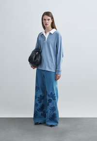 Ανοιχτό μπλε πόλο μπλούζα με μακριά μανίκια και λευκό γιακά· τζιν παντελόνι με φαρδιά μπατζάκια και σκούρα λουλουδάτη κεντημένη διακόσμηση στο κάτω μέρος· μαύρη τσάντα χειρός.