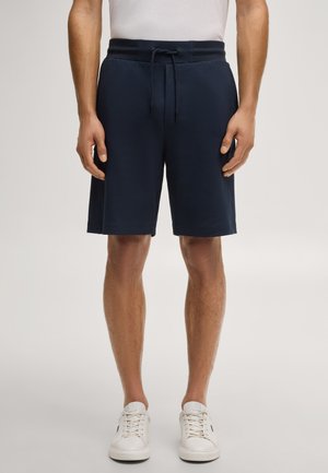 Mężczyzna ubrany w granatowe szorty ze ściągaczem, białe trampki i białą koszulkę, stojący ze spuszczonymi rękami wzdłuż ciała.