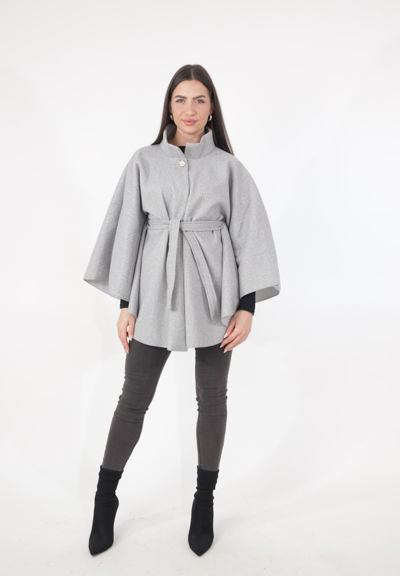 Grijze cape van een wollen blend met een hoge kraag, decoratieve knoop en riem. Heeft brede mouwen en een losse pasvorm, gecombineerd met donkergrijze broek.