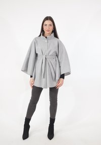 Grijze cape van een wollen blend met een hoge kraag, decoratieve knoop en riem. Heeft brede mouwen en een losse pasvorm, gecombineerd met donkergrijze broek.
