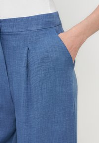 Μπλε υφασμάτινο παντελόνι με πιέτα μπροστά και πλαϊνή τσέπη, συνδυασμένο με λευκό τοπ, το χέρι μέσα στην τσέπη.