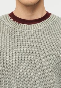 Κοντινό πλάνο ενός ατόμου που φοράει ένα ανοιχτό γκρι πλεκτό πουλόβερ πάνω από ένα μπορντό πουκάμισο με γιακά ριγέ σε λευκό και κόκκινο.