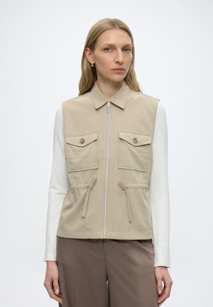 Frau trägt beige ärmellose Weste mit Reißverschluss und zwei mit Knöpfen verschließbaren Brusttaschen über einem weißen Langarmshirt und braunen Hosen, steht vor weißem Hintergrund.