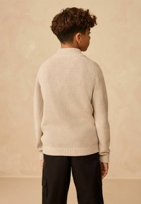 Beiger Strickpullover mit ribbeltem Saum und Bündchen, hochgeschlossen und mit einem strukturierten Muster, kombiniert mit schwarzen Cargo-Hosen.