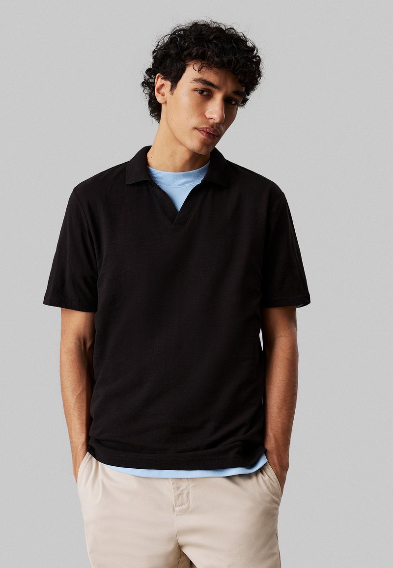 Polo shirt nero a maniche corte con scollatura a V, sovrapposto a una maglietta azzurra a girocollo, indossato con pantaloni beige.