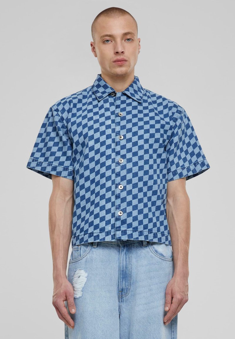 Camicia a maniche corte con motivo a quadretti blu, in materiale denim, con chiusura a bottoni e vestibilità corta e ampia. Pantaloni in denim strappati sotto.