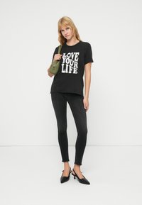 Μαύρο T-shirt με το σχέδιο "LOVE YOUR LIFE", συνδυασμένο με μαύρα στενά τζιν και μαύρα μυτερά τακούνια. Το μοντέλο κρατά μια μικρή πράσινη τσάντα.