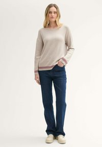 Gebreide trui in lichtbeige met een visgraatpatroon en bordeauxrode afwerking bij de manchetten en de zoom; gecombineerd met blauwe rechte jeans en beige schoenen.