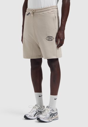 Mann trägt beige Shorts mit Kordelzug und schwarzem Logo, weiße Nike-Socken und silberne Laufschuhe vor einfarbigem Hintergrund.