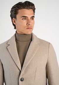 Abrigo de lana beige con solapas y cierre de un solo botón, llevado sobre un suéter de cuello alto marrón texturizado. Tejido suave, ajuste entallado.