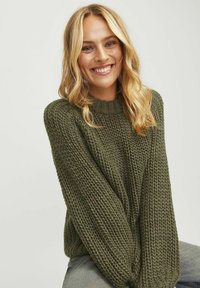 Chunky olijfgroen gebreid sweater met een geribbelde ronde hals, lange mouwen en een gestructureerde stof. Gekozen boven een vervaagde jeans.