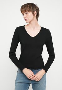 Zwarte langslavertop met geribbelde textuur, V-hals ontwerp en golvende zoomdetail, gedragen met lichtblauwe denim.