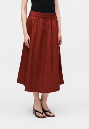 Žena nosiaca medzikrátku hnedočervenú sukňu s elastickým pásom a čierne klinové sandále, stojaca pred jednofarebným pozadím.