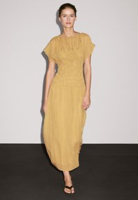 Maxikleid in senfgelb, leichtes Material mit gerafftem Oberteil, kurzen Ärmeln und fließendem Schnitt. Einfache Riemchensandalen.
