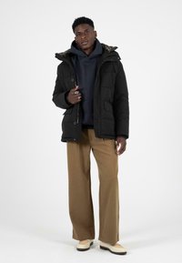 Černá péřová bunda s kapucí podšitou kožešinou, zapínáním na knoflíky a bočními kapsami, v kombinaci s námořnickou mikinou a širokými kalhotami v béžové barvě.