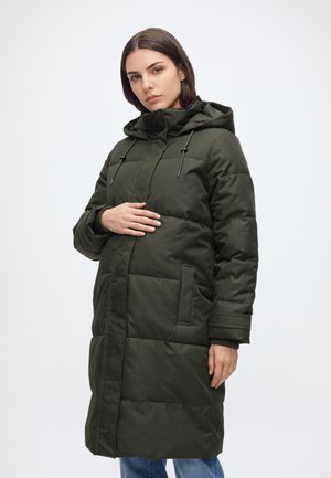 Mujer con abrigo de invierno acolchado verde oscuro con capucha, de pie frente a un fondo liso y claro, sosteniendo su vientre con una mano.