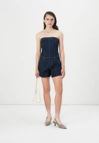Jumpsuit in denim senza spalline con corpetto aderente, gambe corte, tasche laterali e cuciture a contrasto. La modella tiene in mano una borsa bianca.