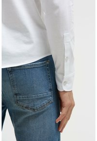 Weißes Langarmshirt mit Knopfmanschetten, kombiniert mit blauen Jeans aus Denim, die eine Gesäßtasche haben. Das Hemd hat eine glatte Textur und eine taillierte Passform.