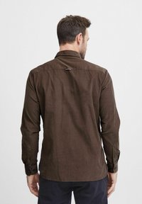 Cămașă maro cu mânecă lungă, cu guler, manșete cu nasturi și detaliu cu buclă la spate. Fabricată dintr-un material texturat, cu un tiv drept.