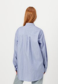 Modra in bela črtasta srajca z natrganjem na hrbtu, dolgimi rokavi in gumbnimi manšetami, narejena iz lahkotnega blaga, z obliko ovratnika.