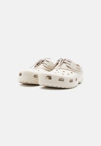 Ζευγάρι μπεζ casual κλογκς με οπές αερισμού, λεπτομέρειες με κορδόνια και υφή στη σόλα, εμφανίζονται από χαμηλή γωνία από μπροστά σε λευκό φόντο.