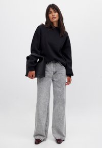 Čierny oversized sveter, šedé široké nohavice a vínové členkové topánky. Model drží čiernu tašku a stojí na jednofarebnom bielom pozadí.