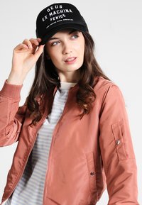 Chaqueta bomber rosa con cremallera en la parte delantera y bolsillos laterales, superpuesta sobre una camiseta de rayas. Gorra negra con logo de texto en blanco.