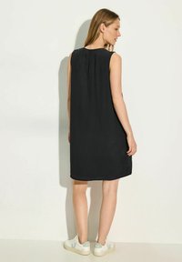 Vestido negro sin mangas de corte holgado, con escote redondeado y tejido suave, combinado con zapatillas blancas con sutiles acentos verdes.
